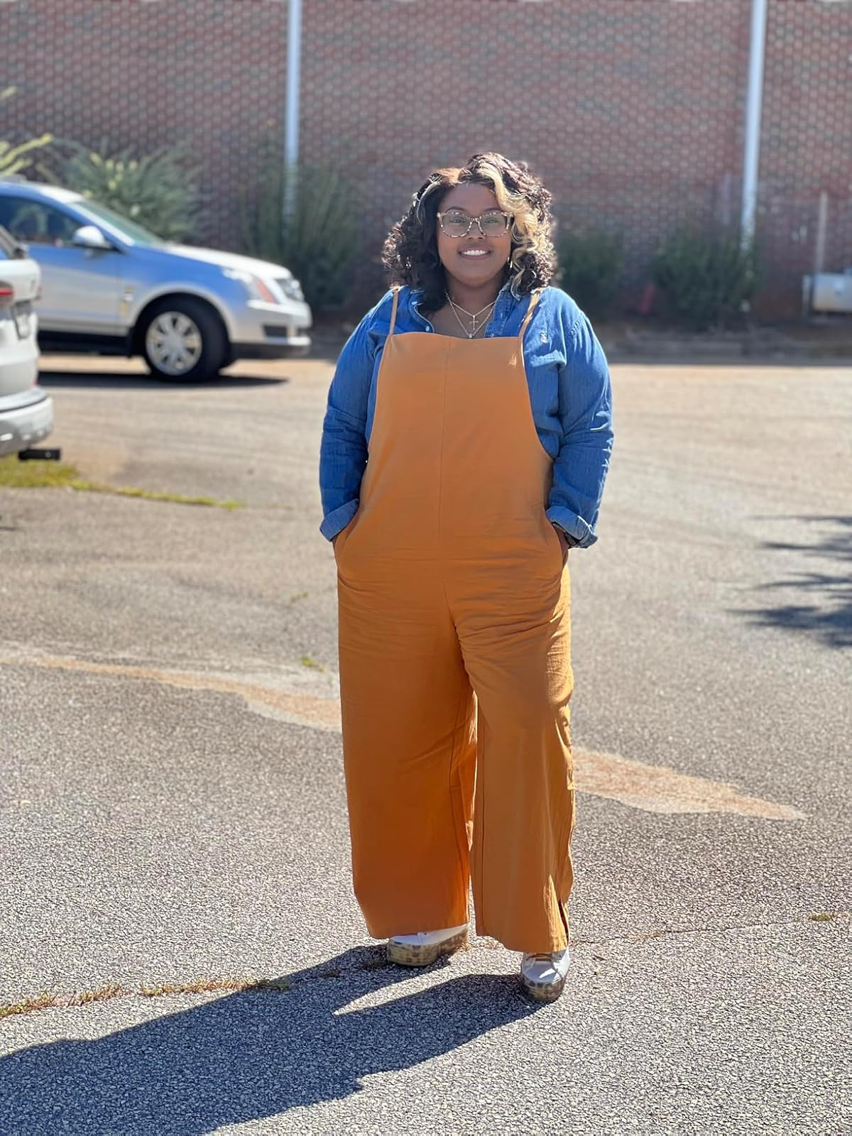 Person in a denim shirt with orange overalls stands in a parking lot, smiling. They wear sunglasses and casual shoes