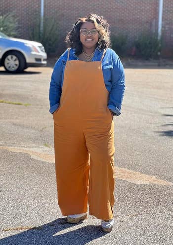 Person in a denim shirt with orange overalls stands in a parking lot, smiling. They wear sunglasses and casual shoes