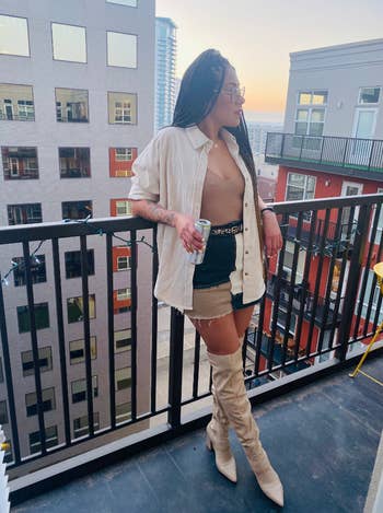 Reviewer on balcony wearing a buttoned shirt, shorts, a belt, and thigh-high boots, holding a drink