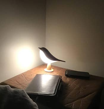 Bird-shaped lamp illuminating a book and phone on a nightstand