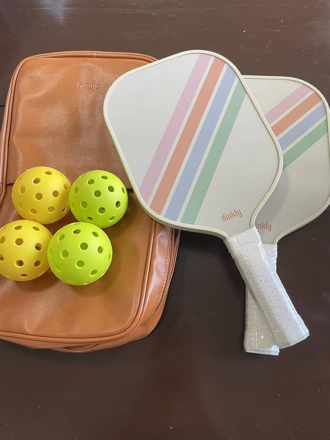 Two pickleball paddles with pastel stripes, a tan bag, and 4  perforated balls connected  a table, showcasing pickleball cogwheel  for sale