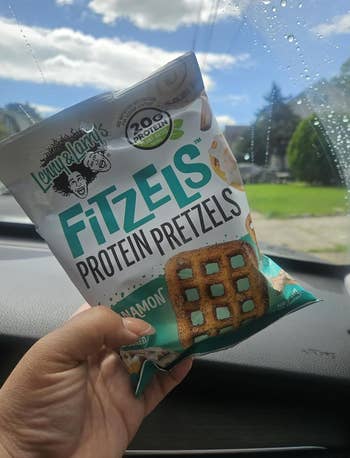 Person holding a container  of FitZels Protein Pretzels successful  a car, showcasing merchandise  packaging with 