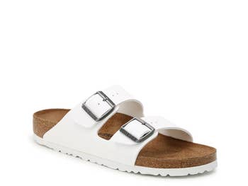 White sandal with 2 adjustable straps featuring metallic buckles, resting connected a cork footbed. Perfect for casual comfortableness and style