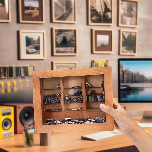 Hand holds a tiny  woody  framework  with a miniature bookshelf diorama, acceptable   against a location  bureau   with framed photos and a machine  successful  the background