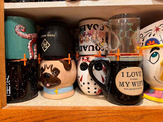 Assorted novelty mugs on a shelf, featuring designs like a pug, masked face, and humorous text, displayed as options for purchase