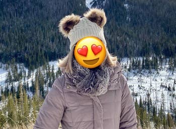 Reviewer wearing white beanie with brown faux fur pom poms on top with a light brown puffer jacket outside