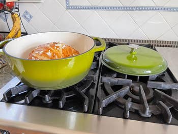 Loaf of bread inside a round, green cast iron pot on a stove. The pot lid is open and resting on another burner