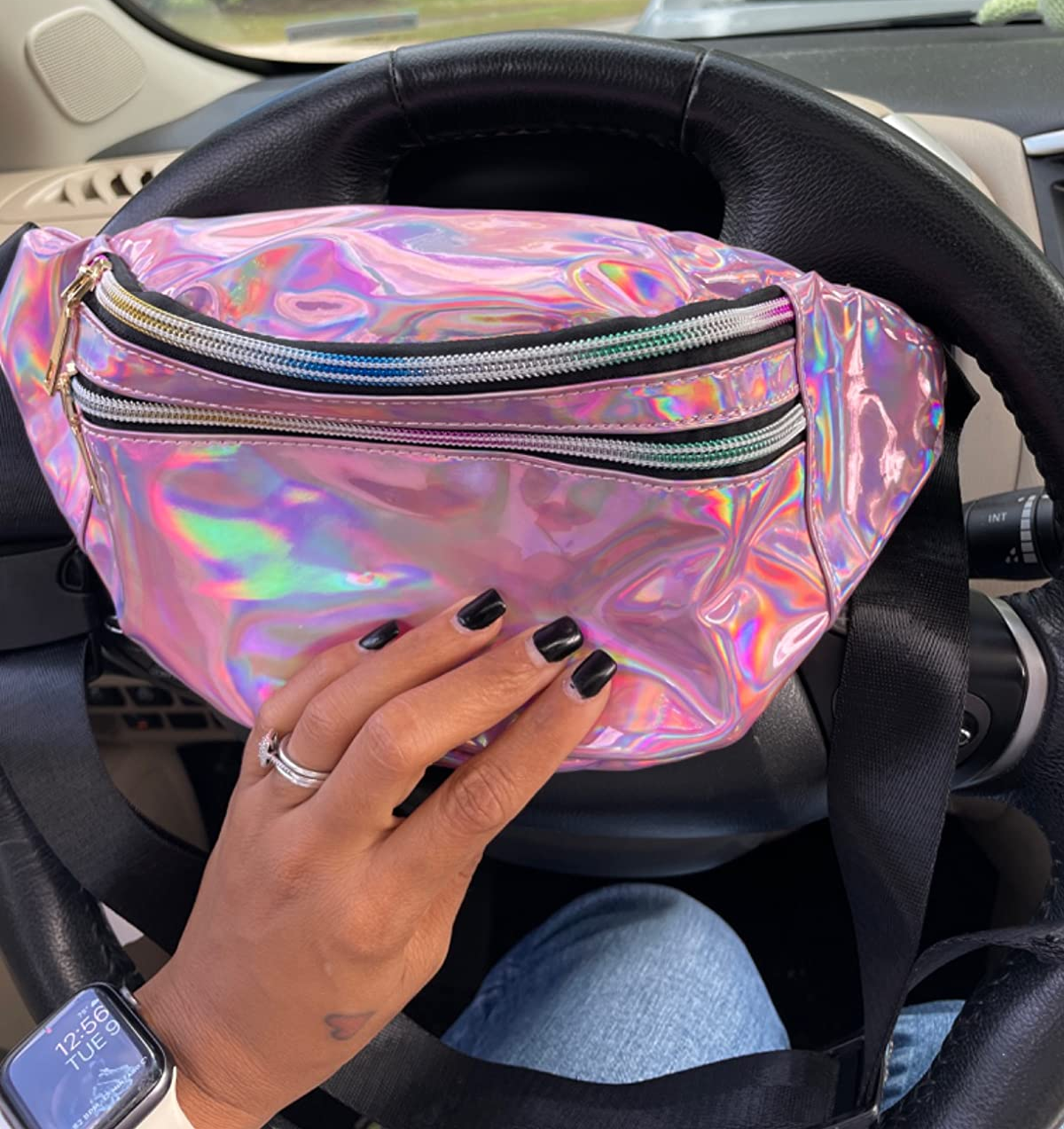 Reviewer holding pink holographic fanny pack with two zipped pouches and a black strap in front of a black steering wheel