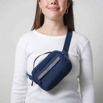 A woman wearing a ribbed long-sleeve top and hoop earrings models a navy crossbody bag with a reflective strip, highlighting a functional and stylish accessory