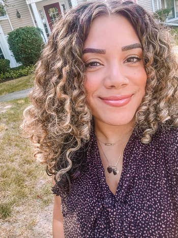 Person with curly hairsbreadth  smiling outdoors, wearing a patterned blouse and layered necklaces, suitable for mundane  manner  inspiration