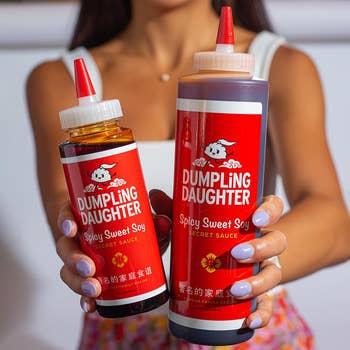 Person holding 2 bottles of Dumpling Daughter Spicy Sweet Soy Secret Sauce, a tiny vessel successful 1 manus and a larger vessel successful the other