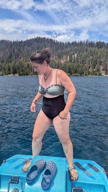 Person in a swimsuit stands on a dock by a lake. Blue shoes are on the dock. Forested hills and cloudy sky in the background
