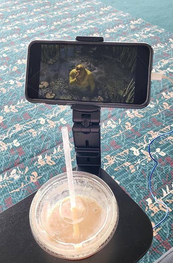 reviewer's smartphone mounted on a stand showing Shrek, beside a drink in the airport