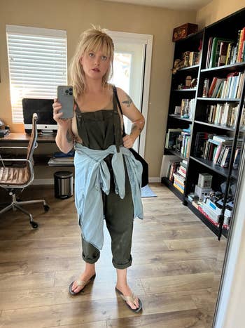 Person successful  an bureau   wearing overalls with a garment  tied astir   the waist, holding a phone, surrounded by bookshelves and a desk. Casual style