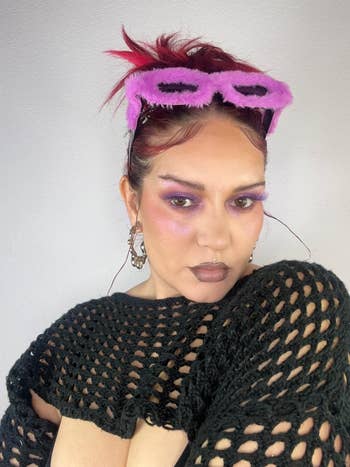 Person wearing a achromatic crochet sweater, bold makeup, and pinkish fuzzy sunglasses connected their head, posing for the camera