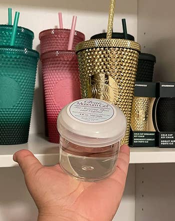 reviewer holding up a jar of museum gel in front of a shelf of colorful drink tumblers
