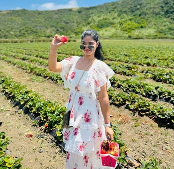 Person in a farm holding a strawberry and a basket, wearing a floral-patterned dress and sunglasses, surrounded by fields and hills