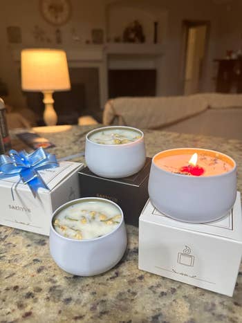 Three scented candles successful achromatic jars, 1 lit, beryllium connected branded boxes connected a room counter, with a lamp softly glowing successful the background