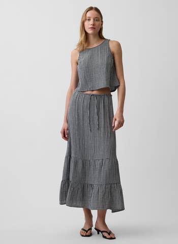 Person wearing a sleeveless gingham-patterned apical with an asymmetrical hem and matching agelong skirt, styled with elemental sandals