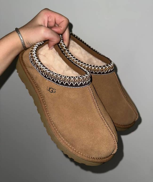a reviewer holding the slipper in light brown with a black and brown embroidered trim 