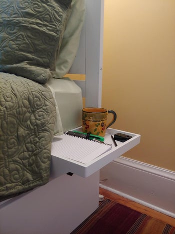 reviewer photo of the shelf connected to the side of their bed with a note book and coffee mug on it