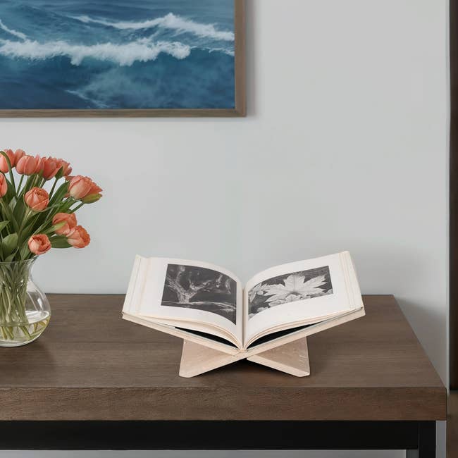 Open book on a wooden stand, displaying black and white images, next to a vase of tulips on a table. Wall art of ocean waves in the background