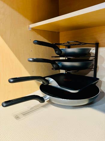 Four frying pans are neatly stacked in a vertical rack inside a wooden kitchen cabinet