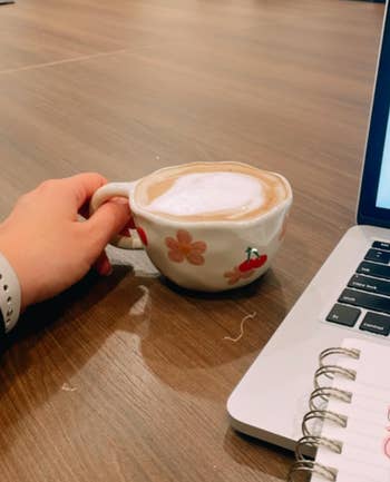 A manus  holding a floral-patterned java  cupful  adjacent  to an unfastened  laptop and a spiral notebook connected  a woody  table