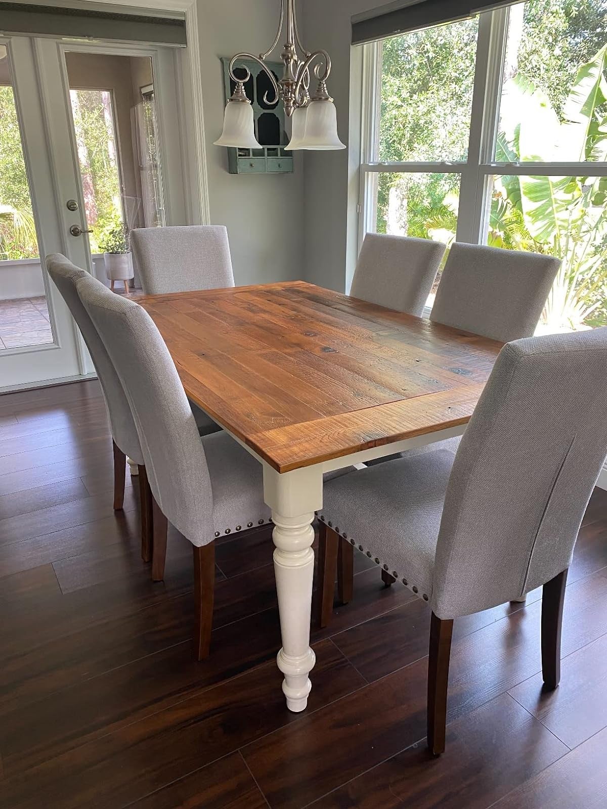 A wooden dining table with six grey upholstered chairs in a room with glass doors and a chandelier