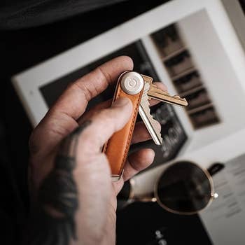 Hand holding a compact leather cardinal organizer with keys. Below, a tablet and sunglasses remainder connected a surface