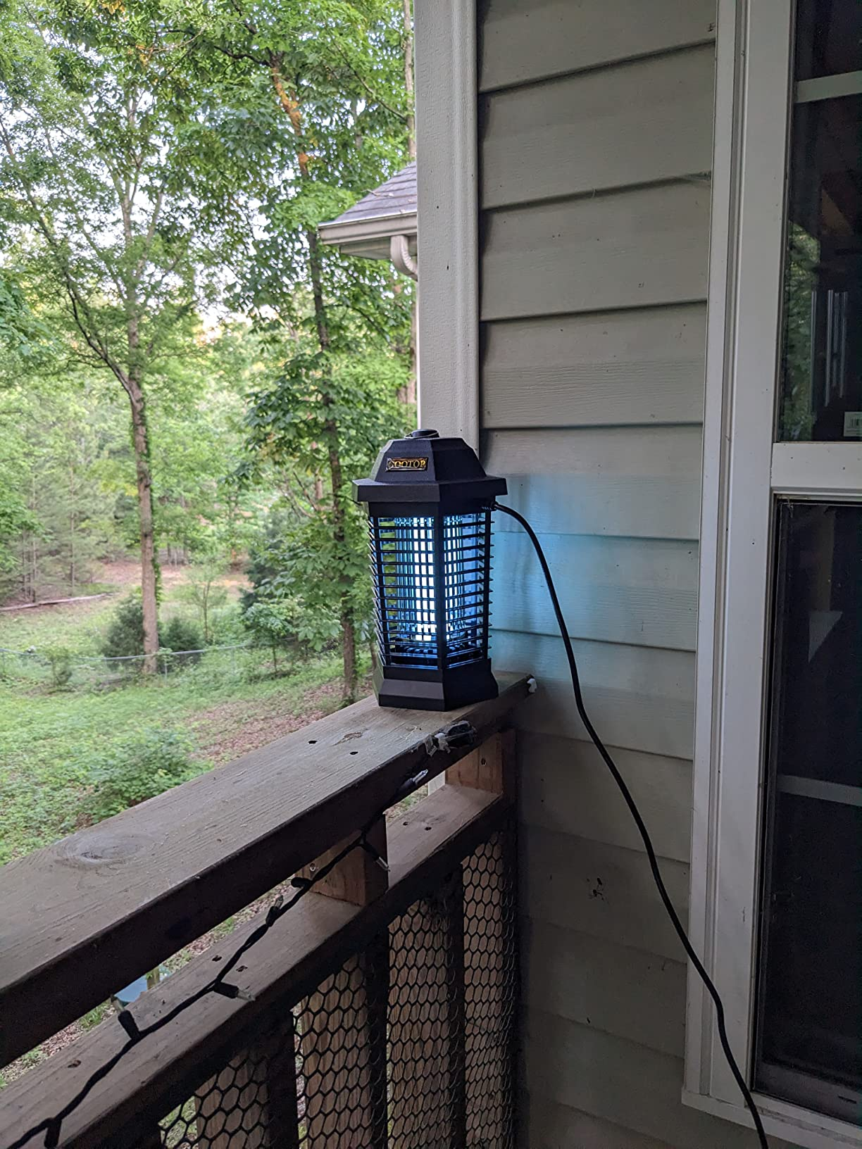 Outdoor electric insect zapper on a wooden porch railing, next to a house wall and window, amidst a background of trees and greenery