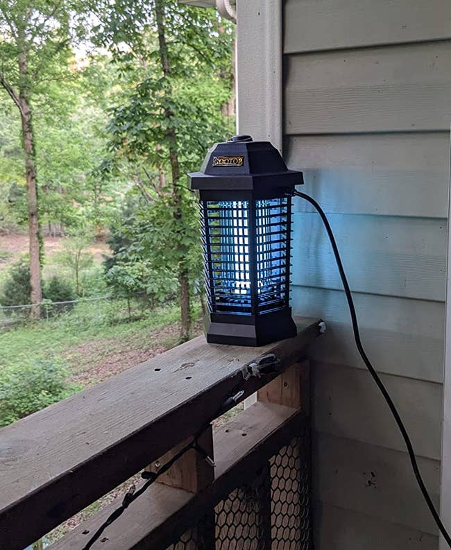 Outdoor electric insect zapper on a wooden porch railing, next to a house wall and window, amidst a background of trees and greenery