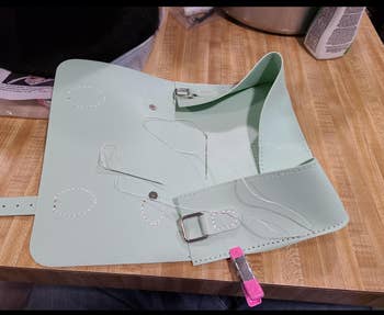 Partially assembled light green leather craft handbag on a wooden table with sewing materials and tools nearby