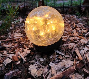 A glowing spherical garden light sits on a bed of wood chips, illuminating its surroundings with a warm light