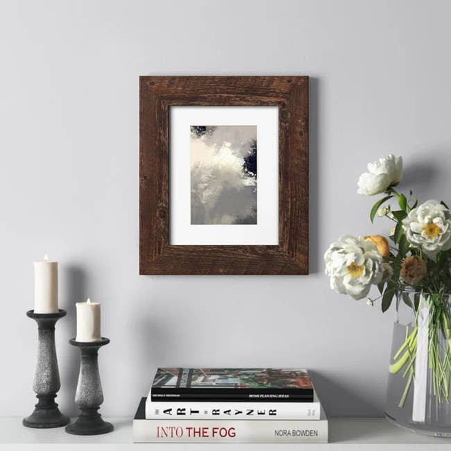 Framed abstract art on a wall beside a stack of books, candles, and white flowers in a vase