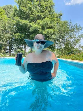Person in a pool wearing a large sun hat and sunglasses, holding a beverage can, with trees in the background