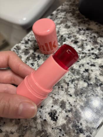 Person holding a red-tinted articulator  balm conduit  with a pinkish  cap, placed connected  a granite countertop