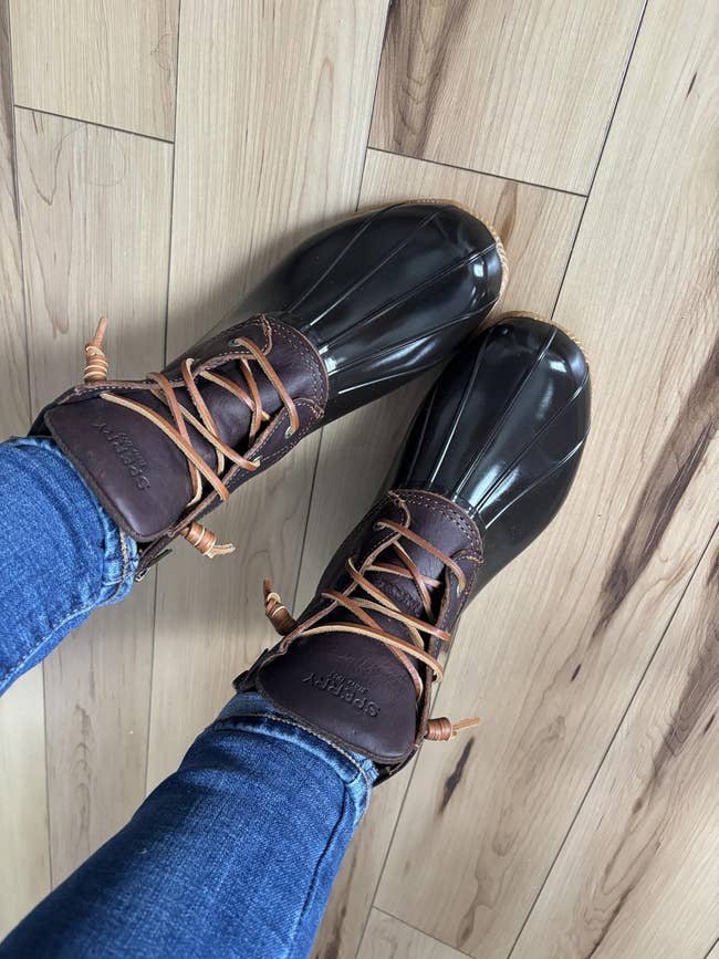 Person wearing duck boots with brownish  leather and rubber soles lasting  connected  wood   flooring