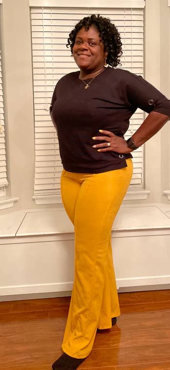 Person smiling, wearing a achromatic apical with fastener details and agleam pants. Standing indoors by achromatic blinds, hands connected hips