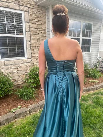A woman stands in a backyard garden wearing a one-shoulder satin gown, styled with an elegant updo. The image showcases the back corseted design of the dress