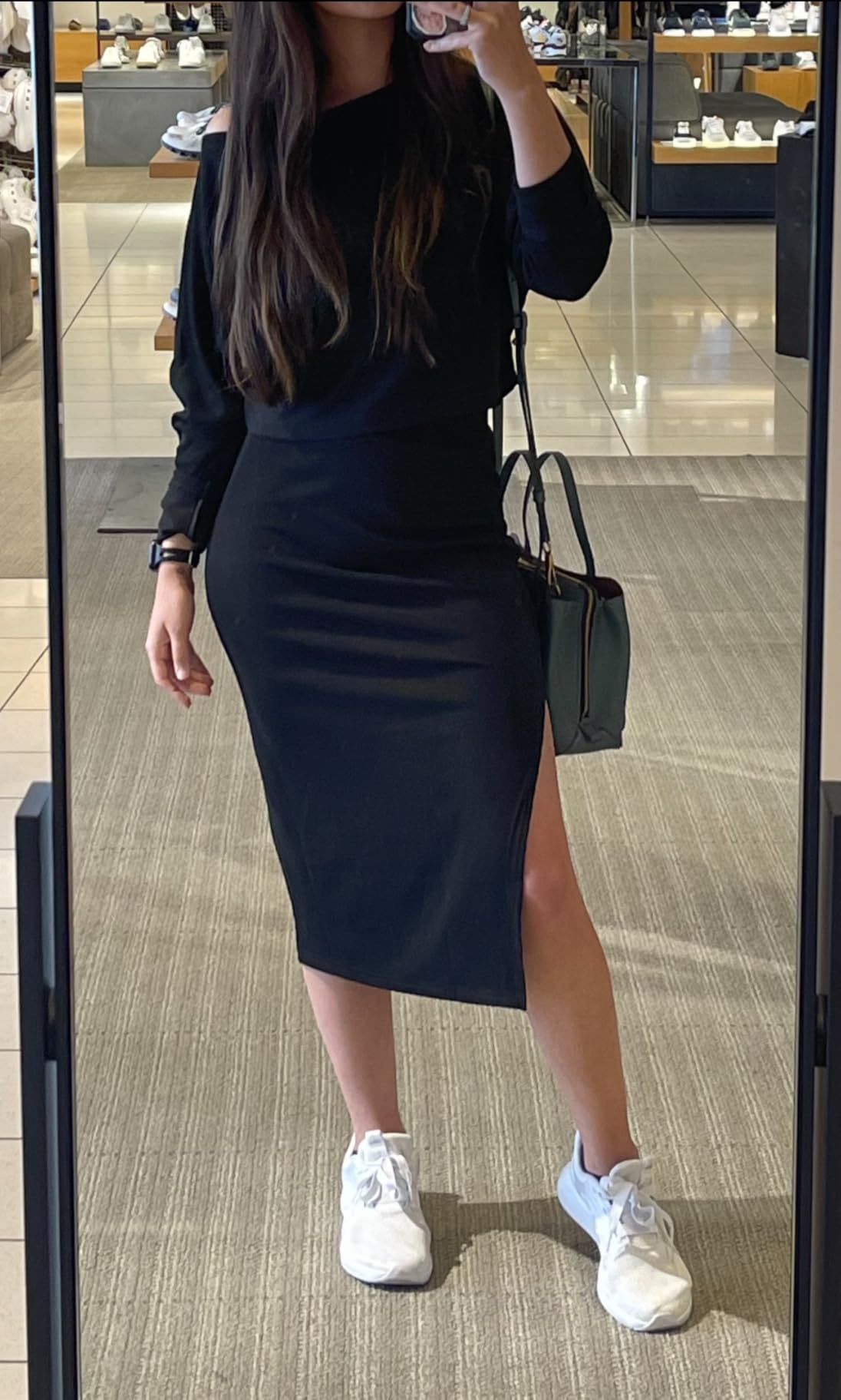 Person in a chic knee-length dress with shoulder cutout, white sneakers, and a handbag, posing in a retail clothing store