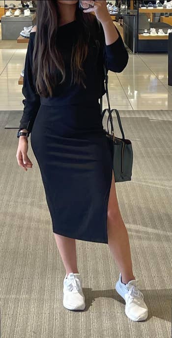 Person in a chic knee-length dress with shoulder cutout, white sneakers, and a handbag, posing in a retail clothing store