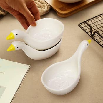 Two ceramic duck-shaped bowls stacked on a table, with one bowl being picked up by a hand. A wire rack and a book are nearby