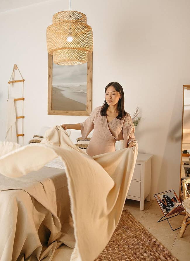 Woman in a cozy room fluffing a beige blanket, wearing casual attire, near a wooden ladder and mirror