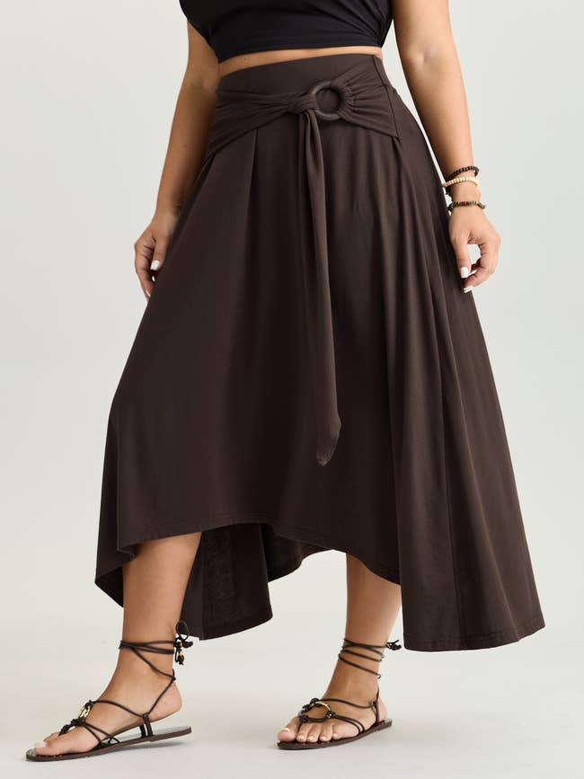 Woman modeling an asymmetrical midi skirt with a beforehand   necktie  and ringing  detail, wearing a achromatic  apical  and strappy sandals