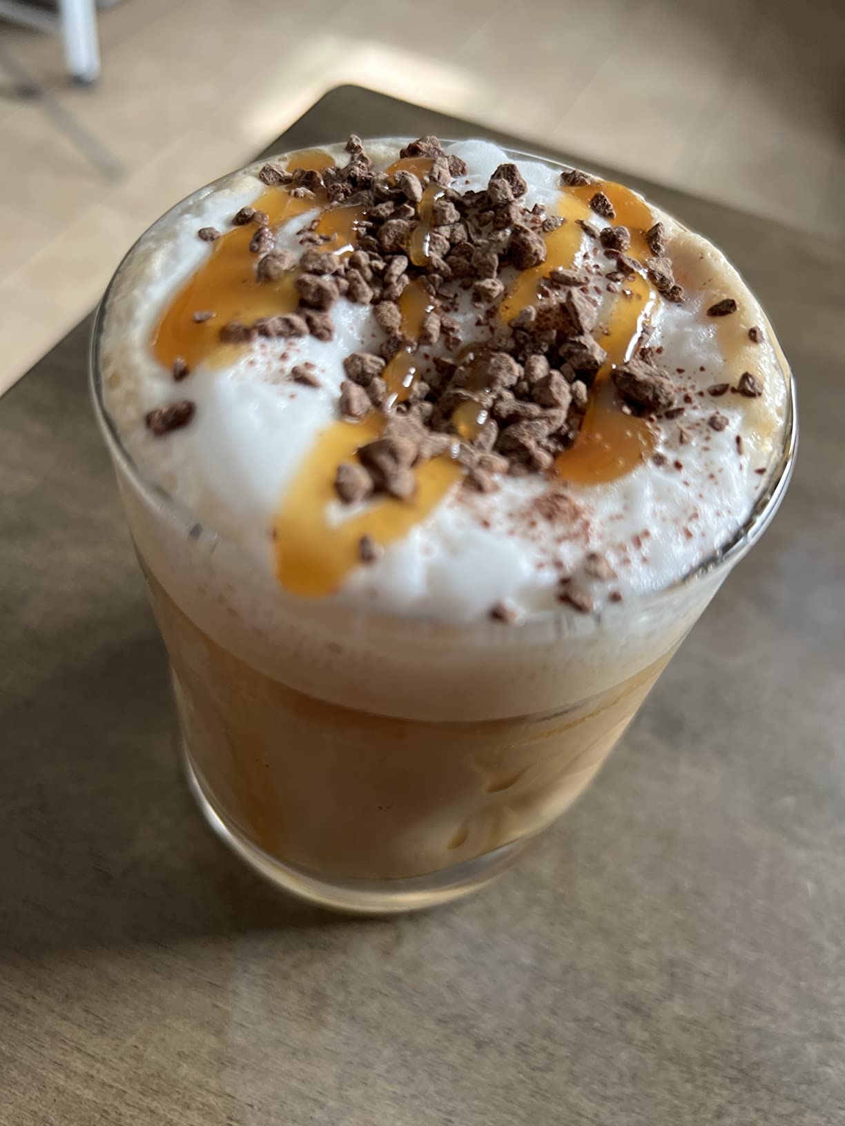 Glass of iced java  topped with frothy cream, caramel drizzle, and cocoa  shavings, sitting connected  a table