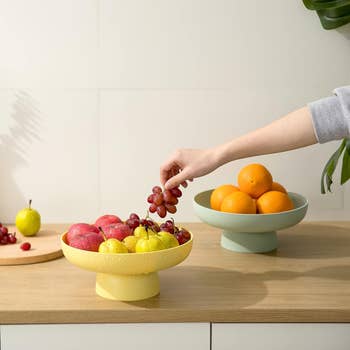 A hand reaches to pick grapes from a bowl of apples and pears, with a bowl of oranges in the background on a modern kitchen counter