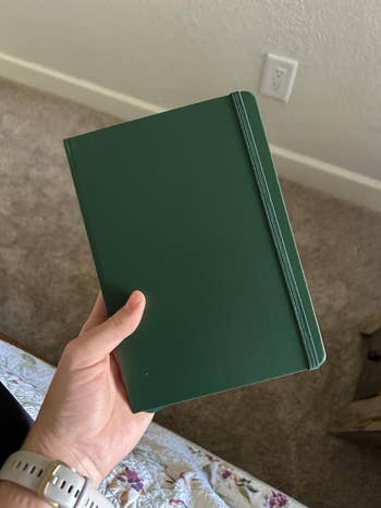 Person holding a closed greenish notebook with an elastic band, lasting successful a carpeted room