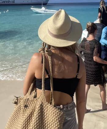 Person successful  a straw chapeau  and achromatic  swimsuit connected  a beach, holding a textured bag. Boat and water  successful  background