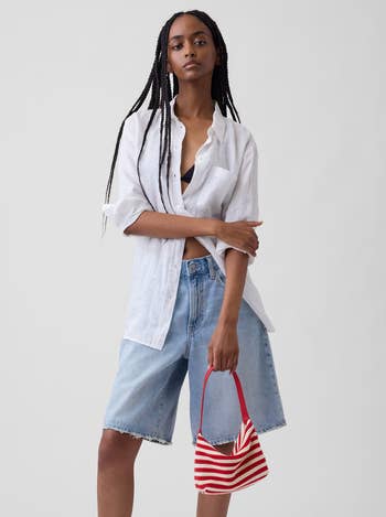 Model successful  a achromatic  button-up garment  and denim shorts holds a tiny  striped bag, showcasing a casual manner  look